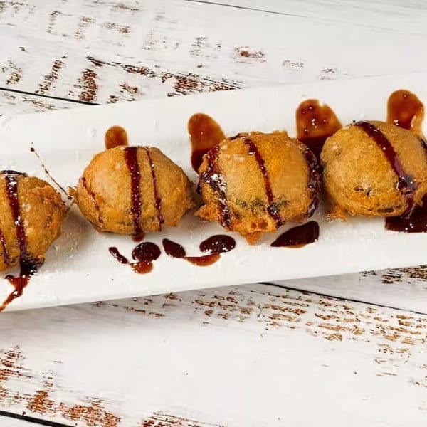Four golden-brown fried dessert balls drizzled with dark syrup and powdered sugar on a white plate.